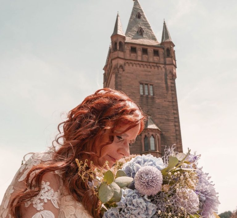Hochzeit Weinheim Braut mit rotem Haar hält einen Blumenstrauß vor einem historischen Gebäude.